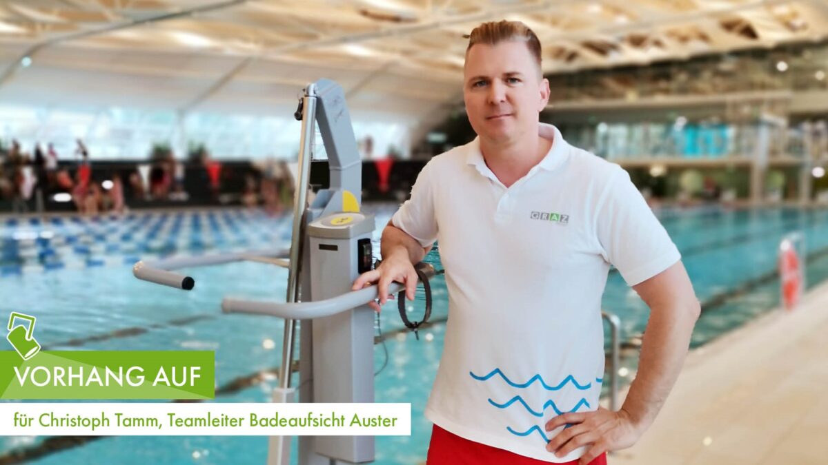 Ein Teamleiter der Badeaufsicht steht lächelnd vor einem Schwimmbecken in moderner Schwimmhalle.